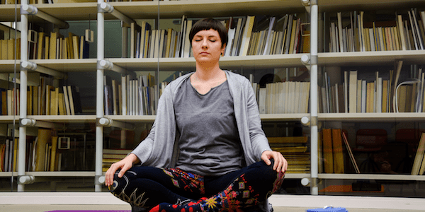 A woman sitting on a yoga mat in a library studying for a master's in yoga therapy.